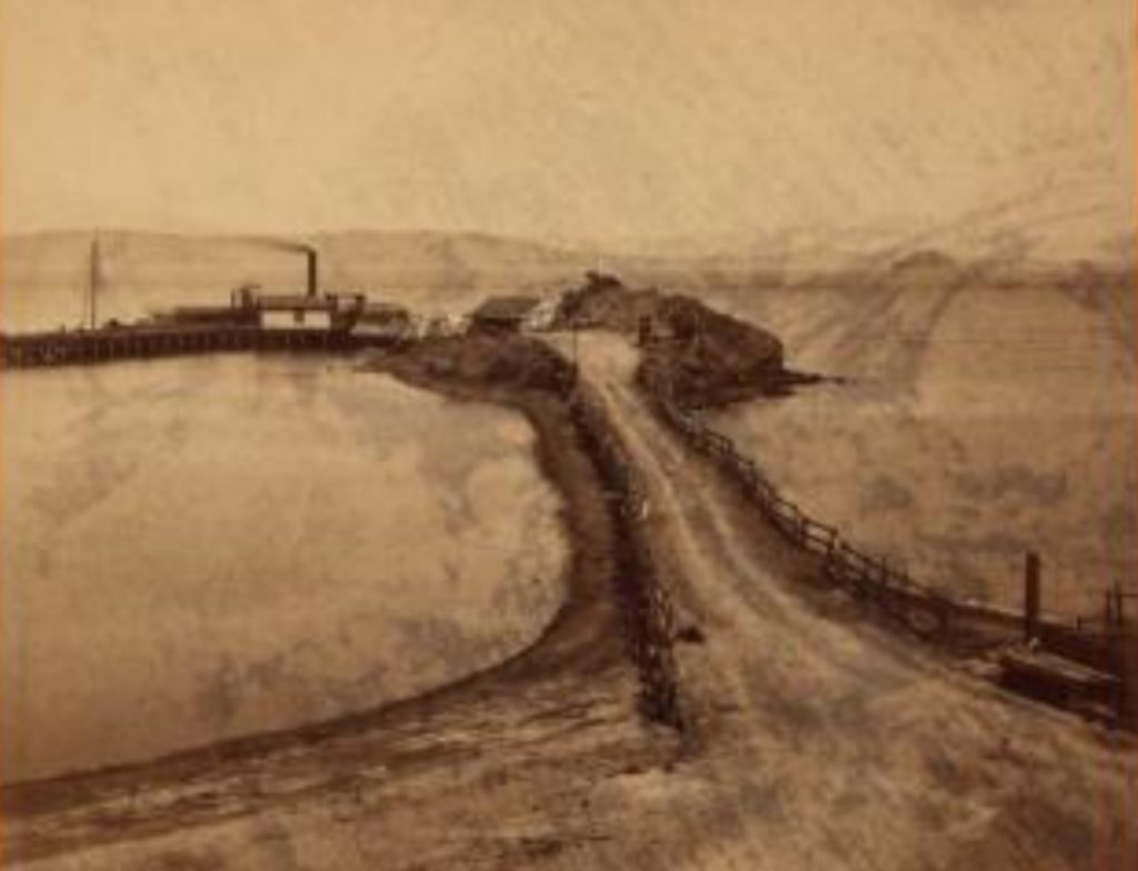 A dirt road leads into a cove where a steamship landing is seen in the distance.