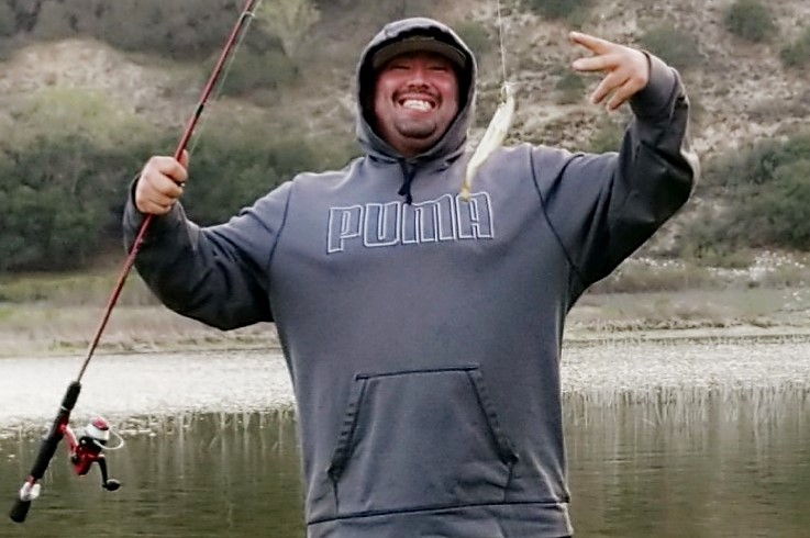 Man wearing hoodie poses with his fishing pole and a small fish.