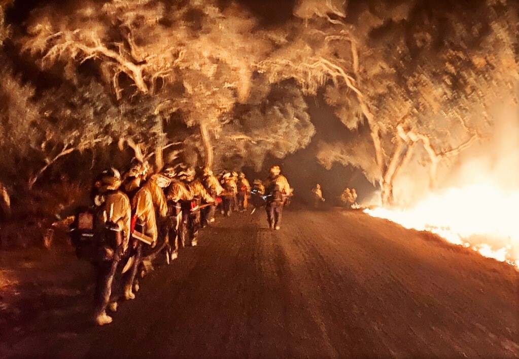Firefighters walk along a dirt road while a fire burns on one side.