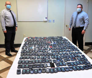 Two men in masks flanking a table with cell phones lined up in rows.