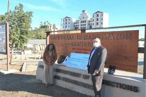 Woman and man wear masks and stand beside the California Rehabilitation Center sign.