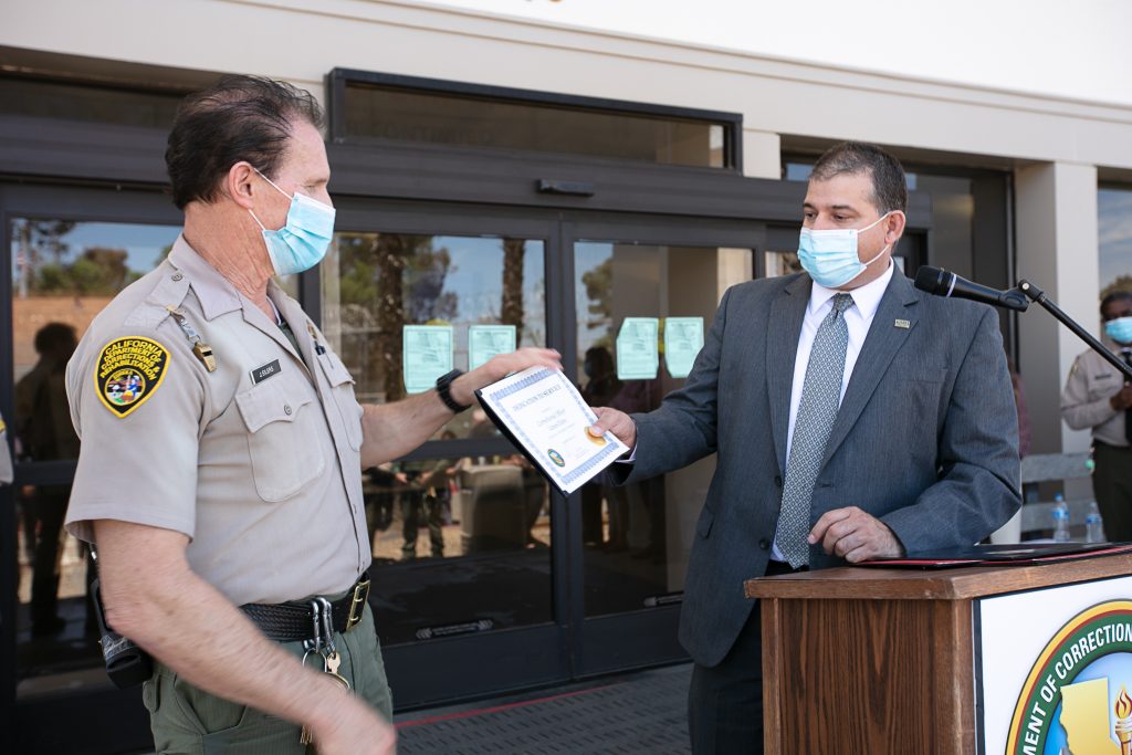 Man in uniform receiving certificate from man in suit with both wearing masks.