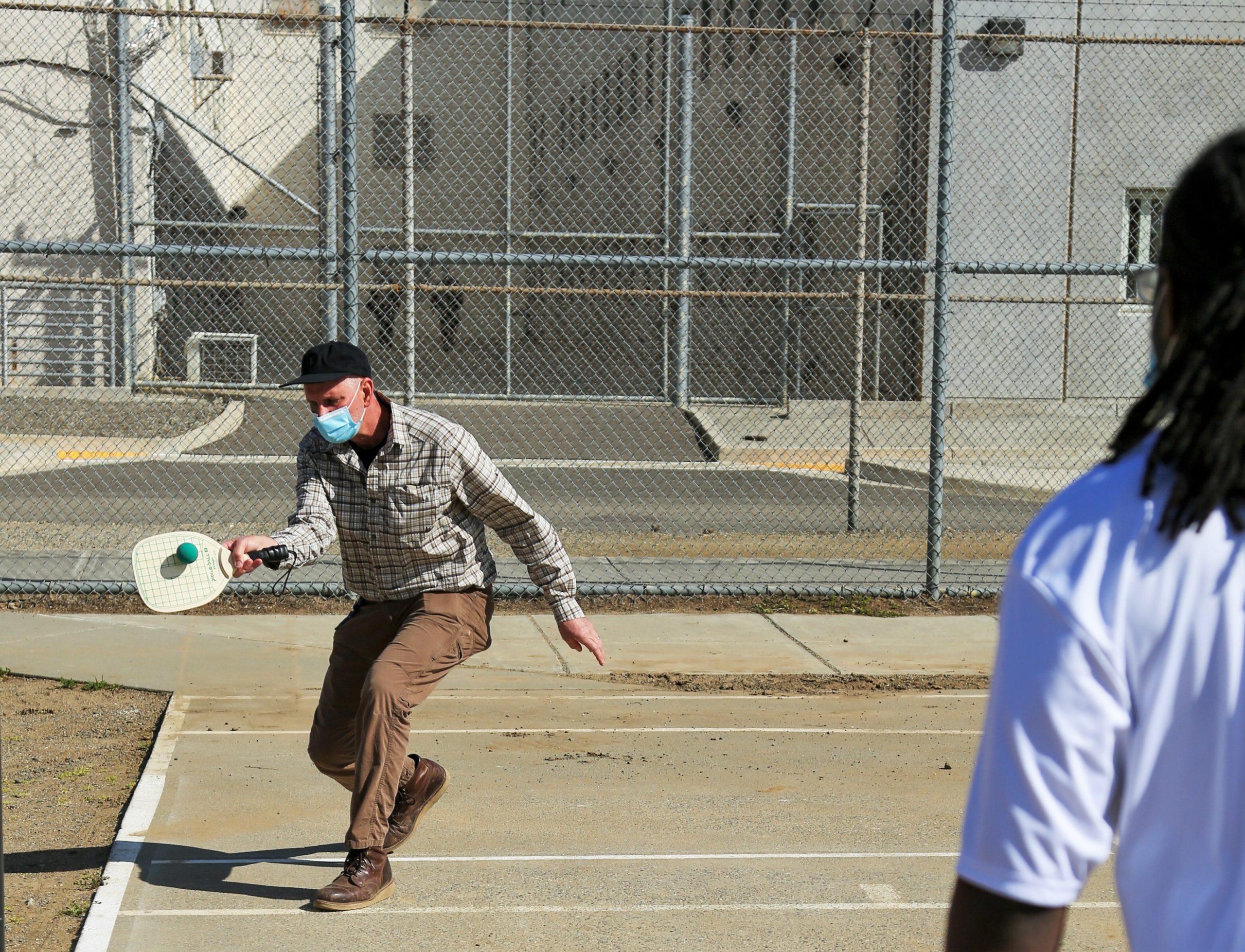 A recreation supervisor plays tennis at CMF, fencing and prison buildings can be seen in the background.