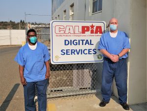 Two incarcerated men stand beside a CALPIA digital services sign.