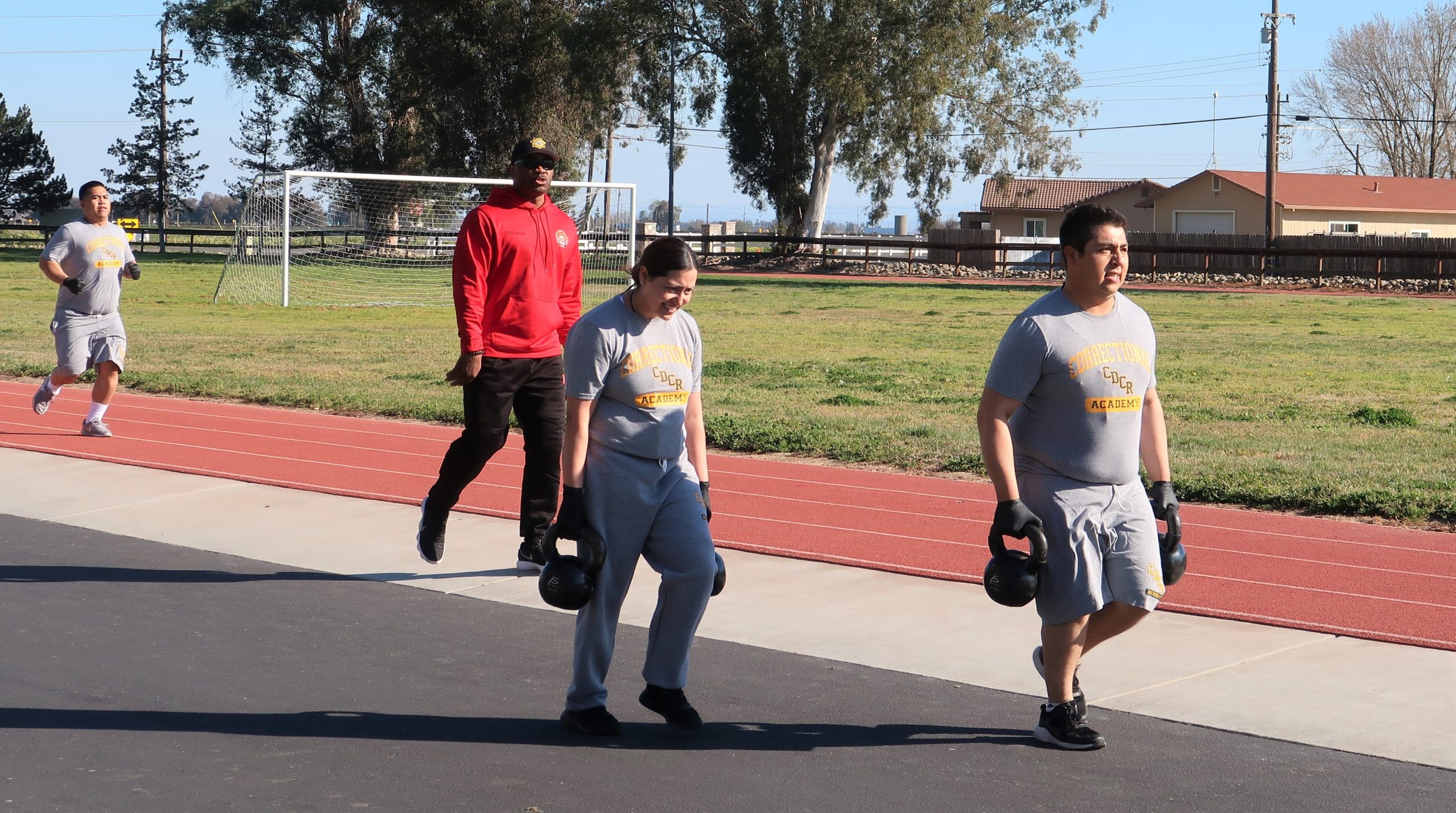 Two cadets carry weights and others run a track while a sergeant watches.