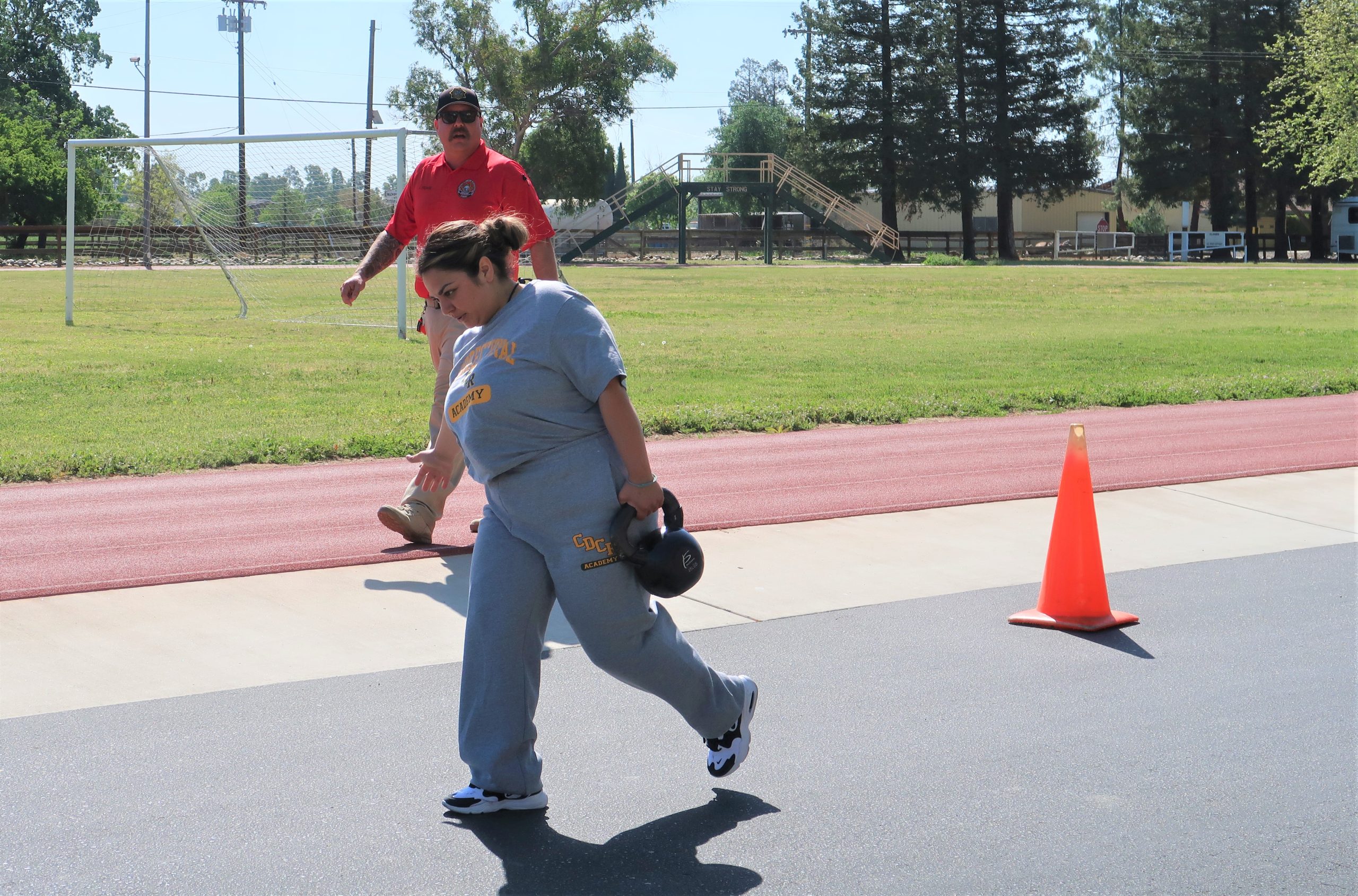 Cadet carries a weight while a sergeant encourages her from the sidelines.