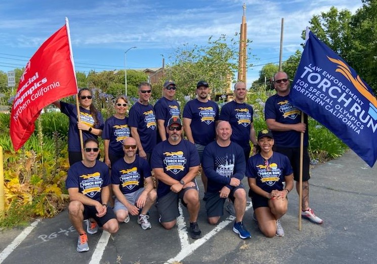 A team of people wearing matching shirts hold flags representing Special Olympics and the Law Enforcement Torch Run.