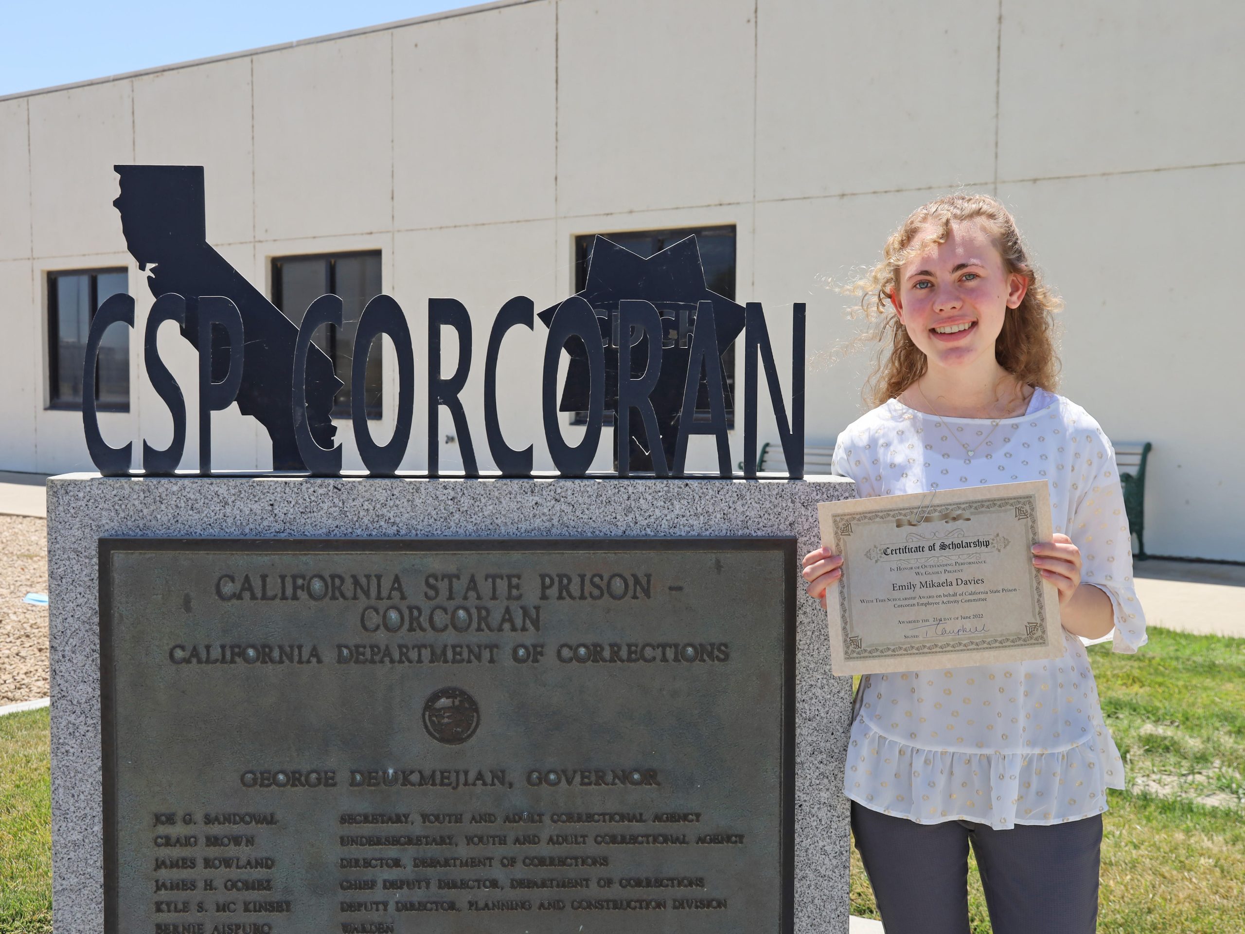 Young woman holds certificate beside a sign for CSP-Corcoran.