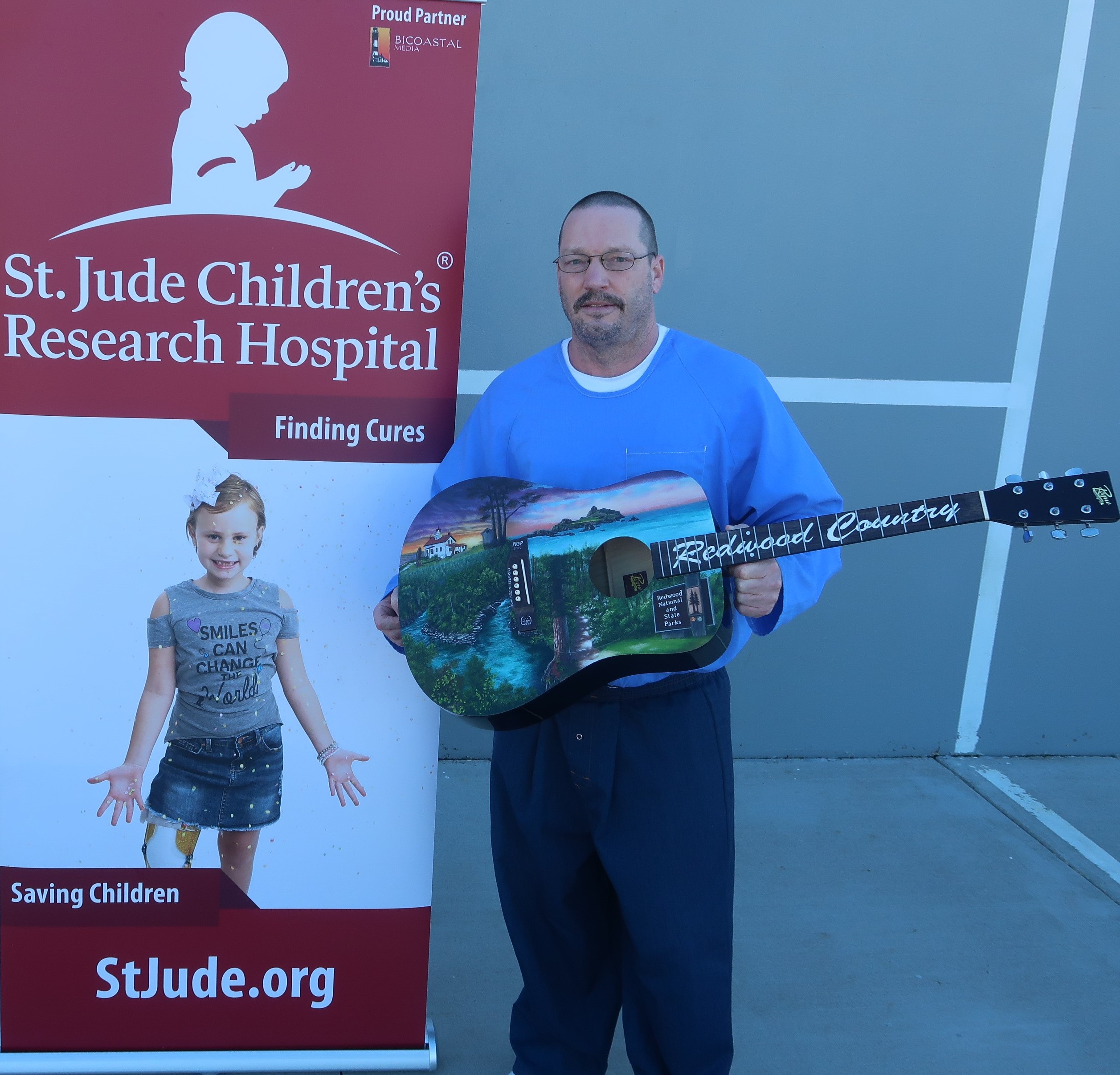 An incarcerated person holds up a painted guitar featuring redwoods and the ocean