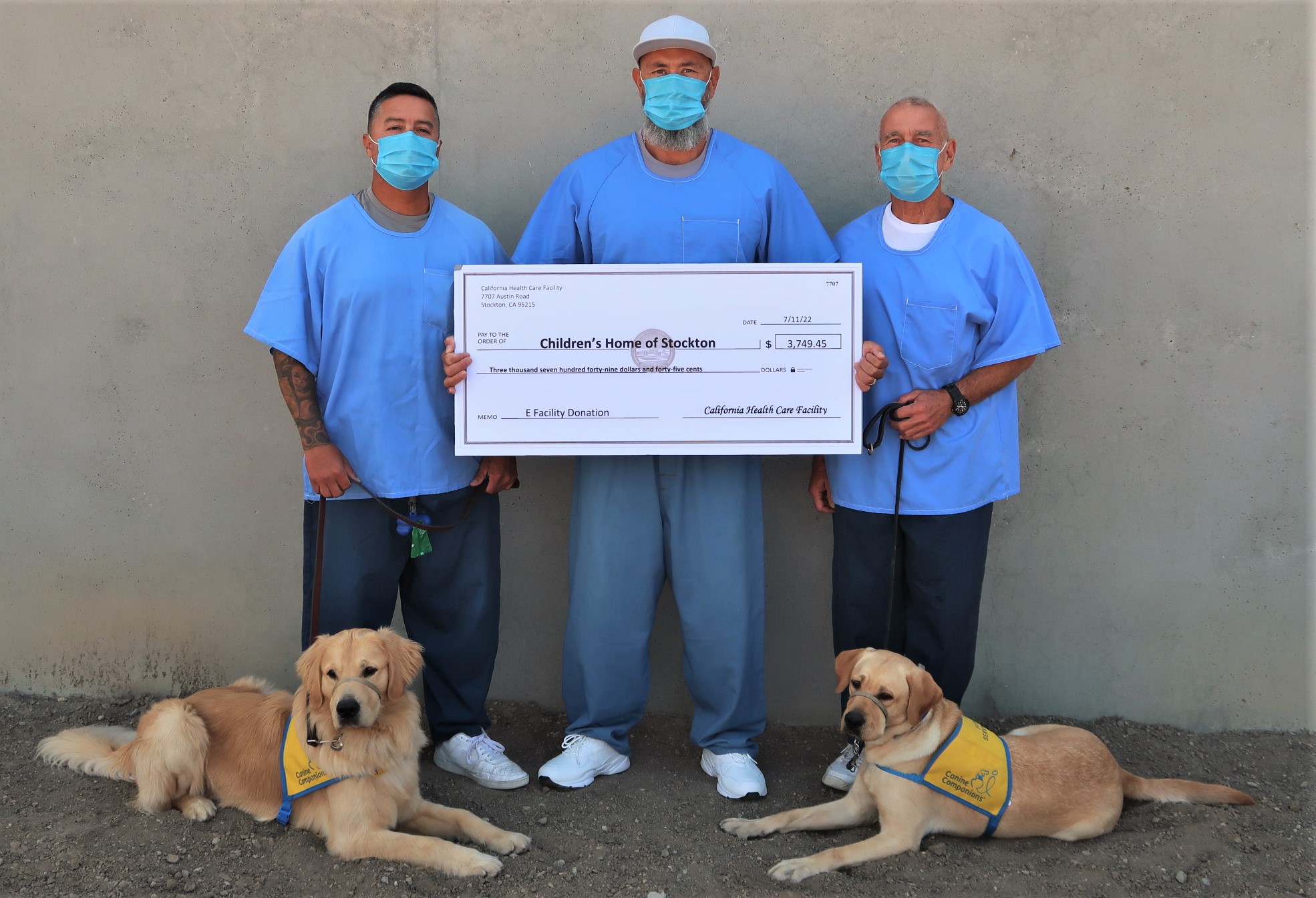 Three incarcerated men hold an oversized check while two dogs rest at their feet.