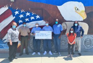 Incarcerated men hold a check for Uvalde school shooting victims while officers and others stand nearby, in front of a giant bald eagle and flag mural.