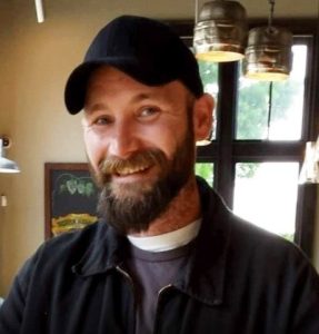 Bearded man wearing hat and jacket smiling.