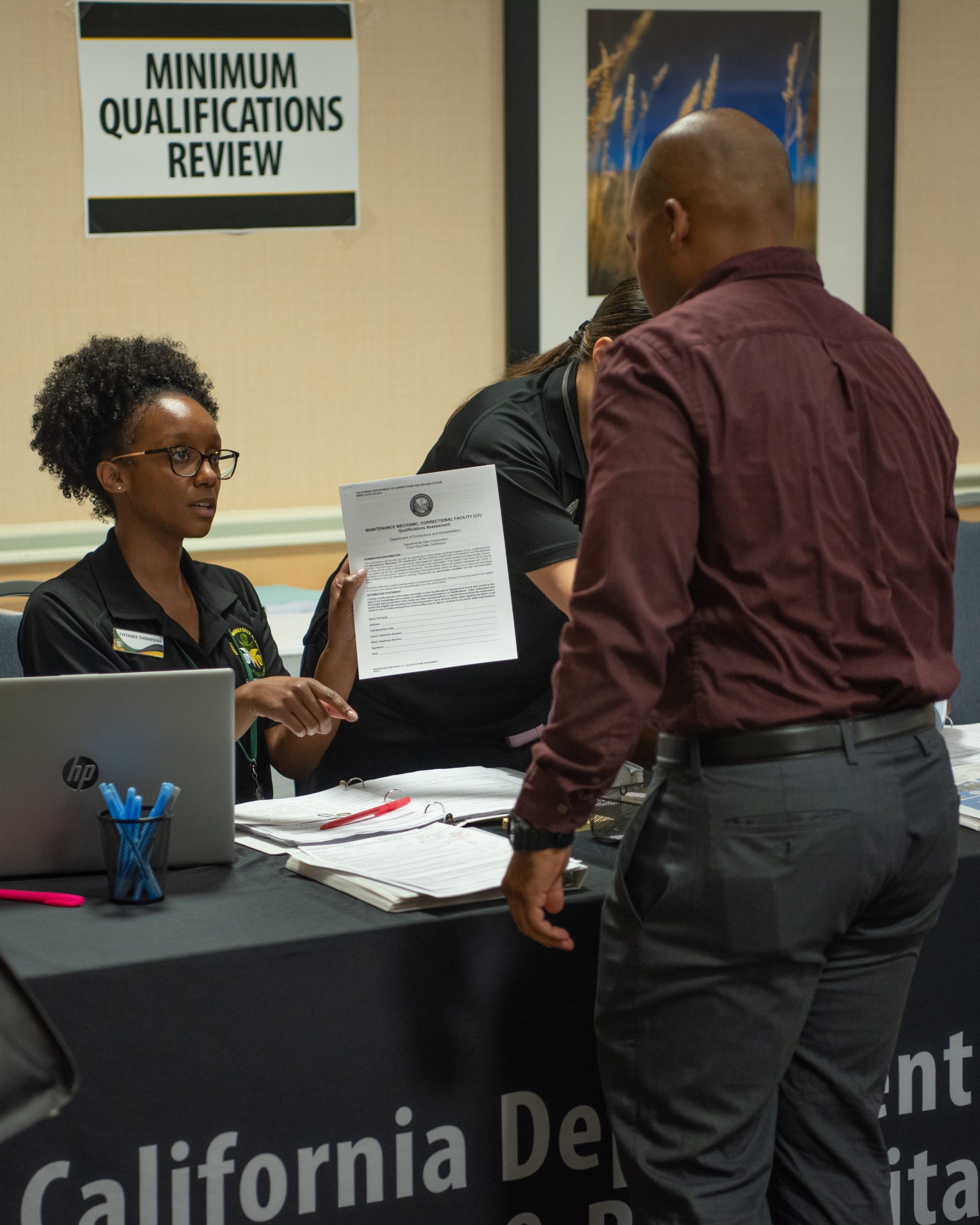 A woman holding a piece of paper speaks to a man at a booth during a hiring event.
