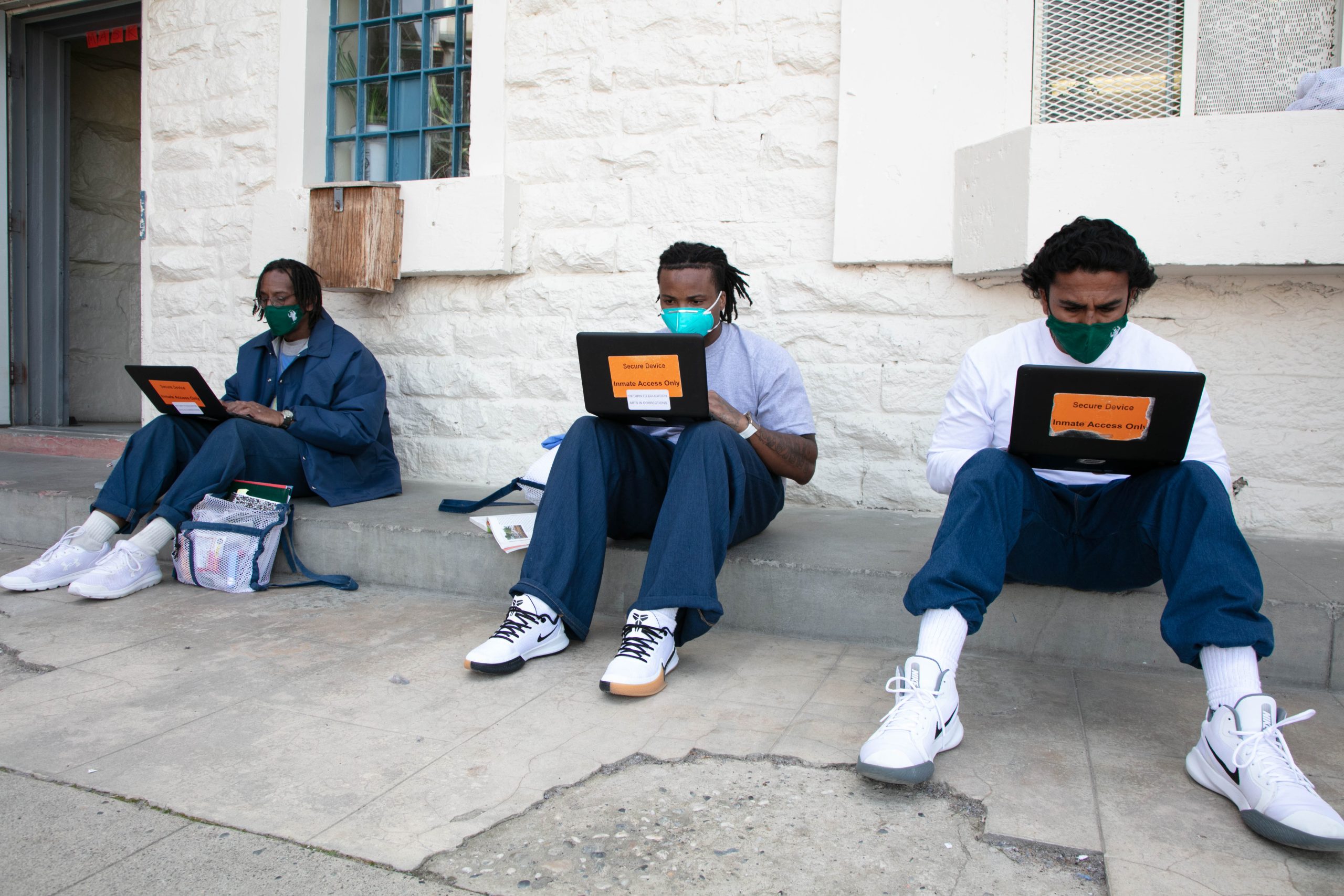 Three incarcerated men sit against a brick wall and use laptops.