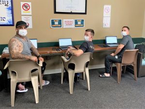 Three men wear masks while sitting in front of laptop computers.