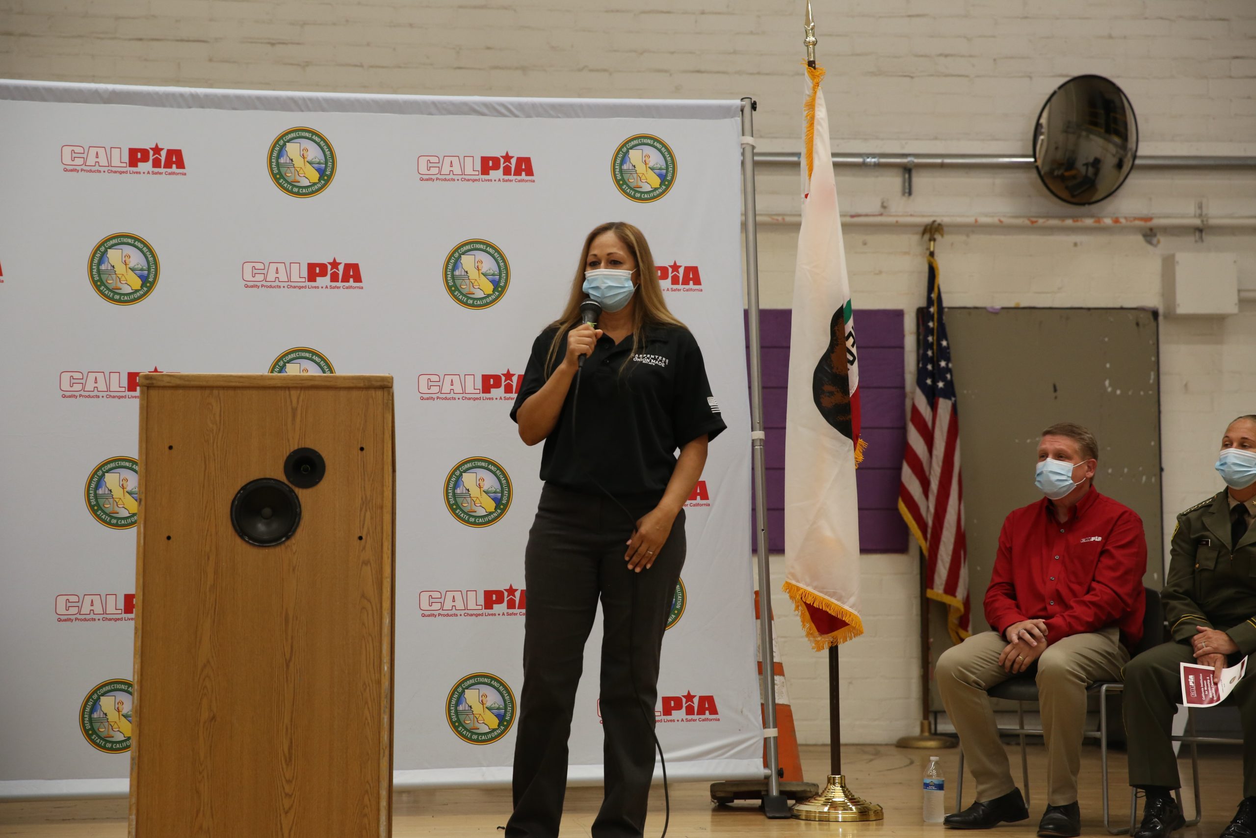 A woman in a mask and black polo shirt speaks near a lectern.
