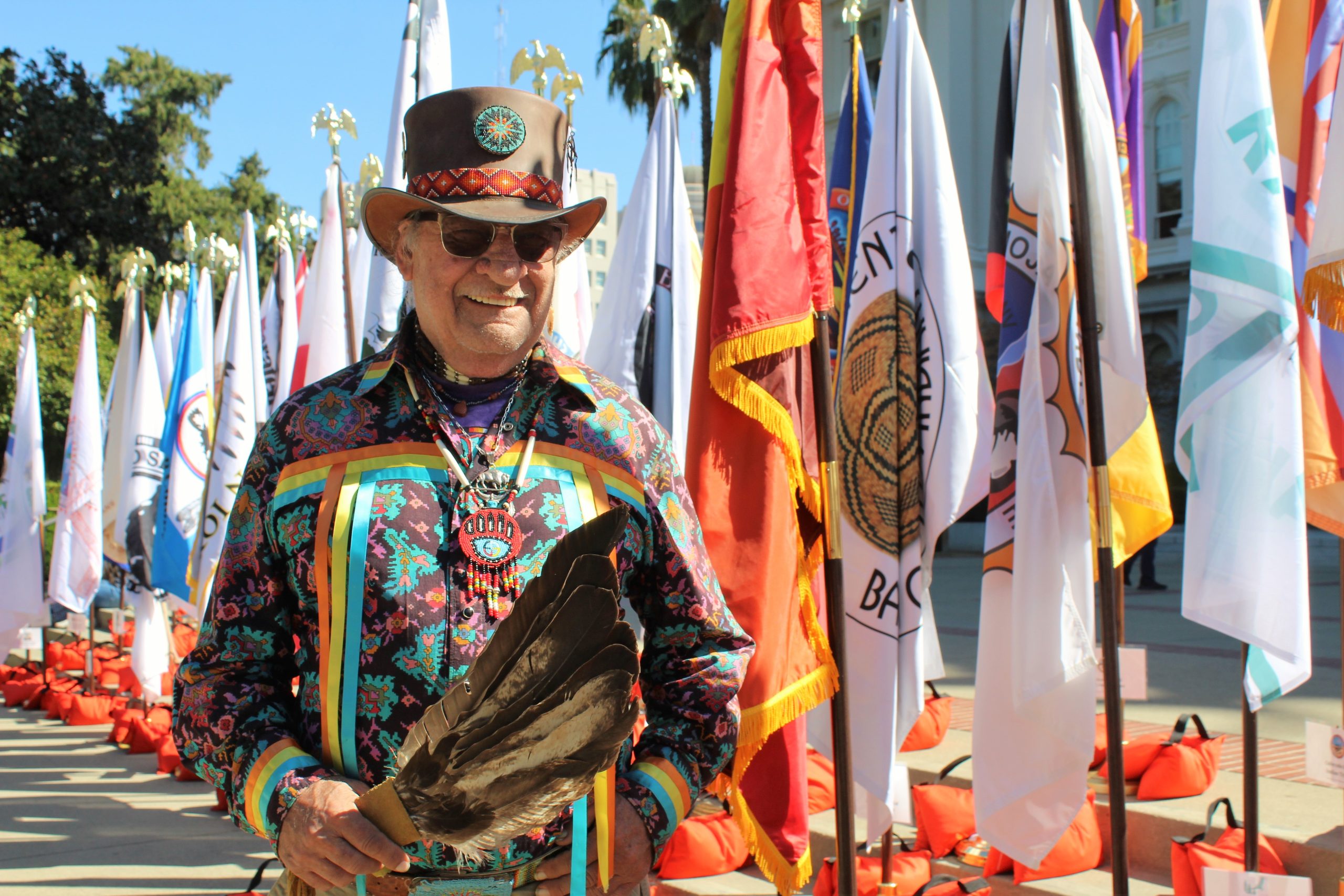 Man in front of Native American tribal flags while holding feathers.