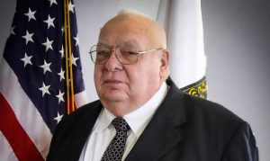 Man wearing glasses, coat and tie in front of flags.
