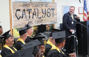People wearing caps and gowns while man speaks into microphone in front of banner that reads OMCP Class of 22 Catalyst CMC Cohort 1.