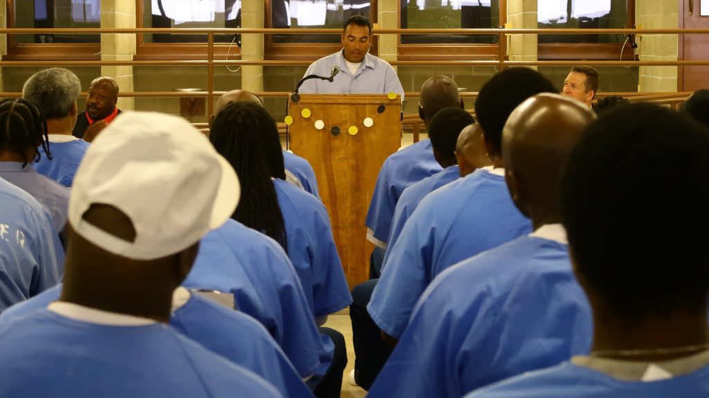 Man at lectern speaks to others at CDCR rehabilitation graduation ceremony.