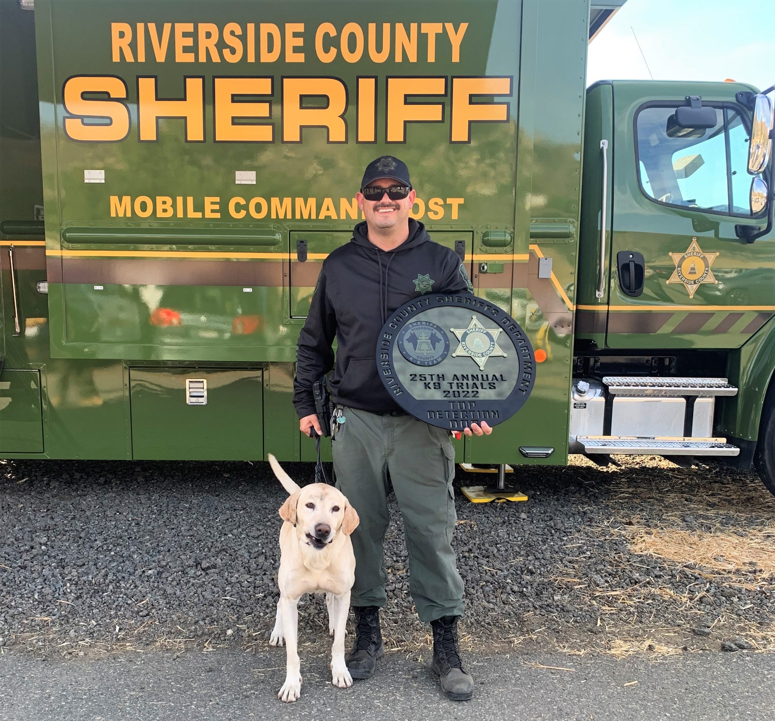 CDCR Officer and K-9 Camo hold award while standing in front of Riverside County Sheriff Mobile Command Unit.