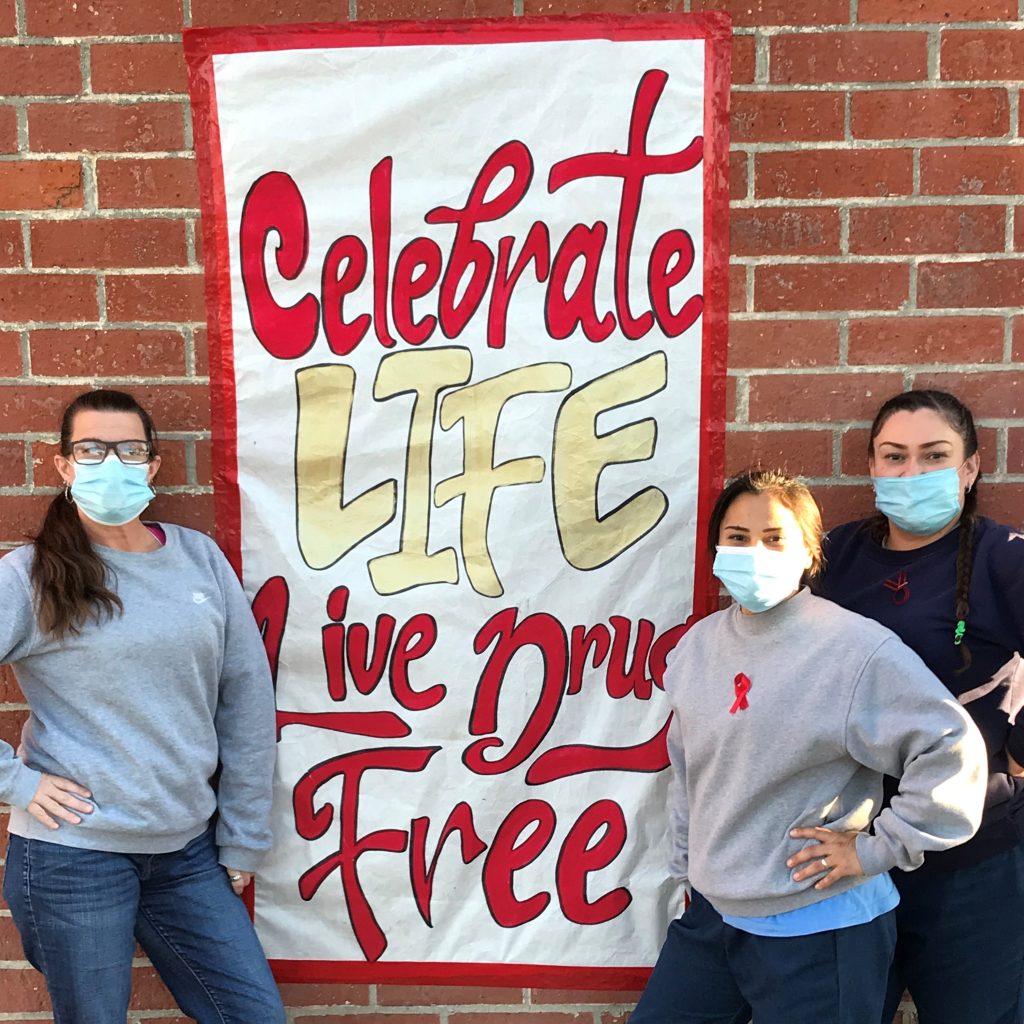 Red Ribbon Week poster at CIW says "celebrate life, live drug free" while three people stand in front of it.