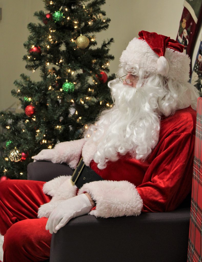 Man in Santa outfit sits beside a Christmas tree.