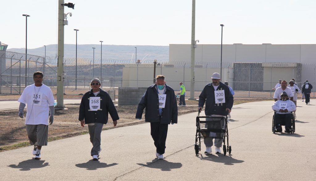 Incarcerated people walking, pushing a walker or in a wheelchair move along a track in a prison.