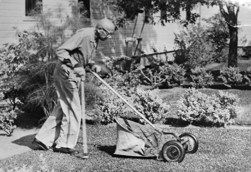 Older man using a cane pushes a non-motorized lawn mower.