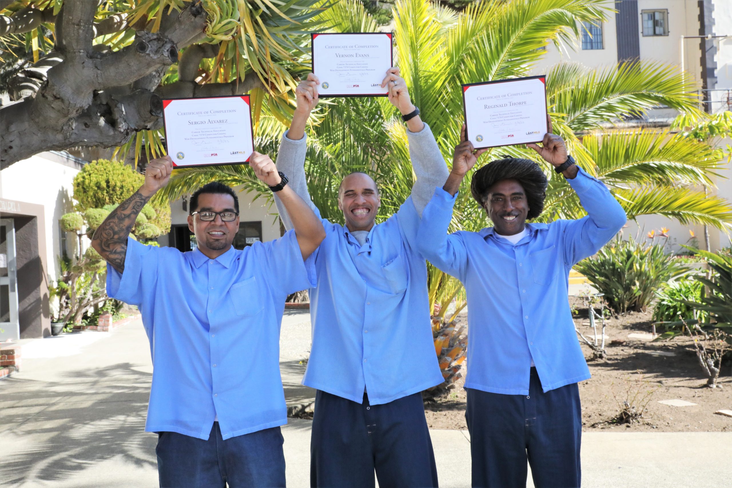 Three incarcerated people hold certificates over their heads.