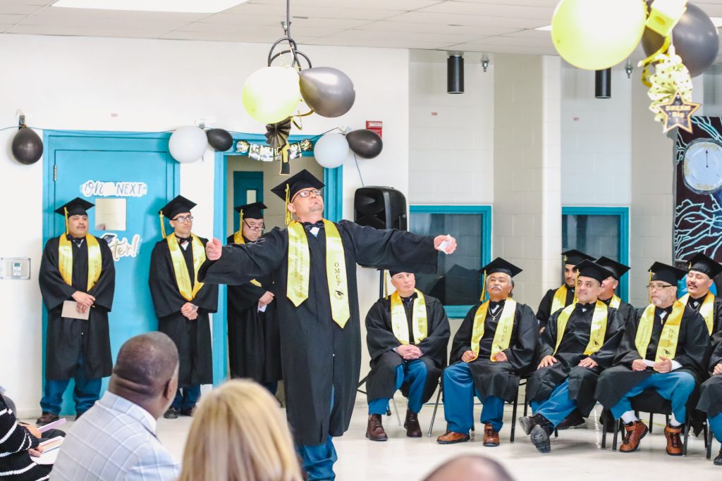 A mentor certification graduate wearing cap and gown walks with his arms outstretched.