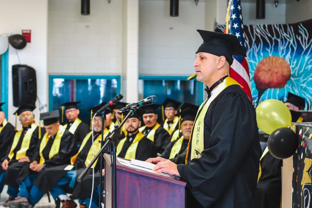 A man speaks at a lectern during a graduation ceremony inside a prison.