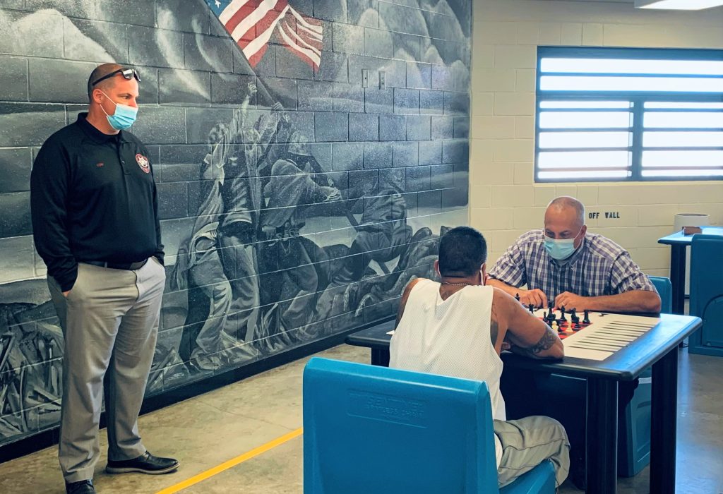 A man in a black polo watches another man and an incarcerated person play chess.