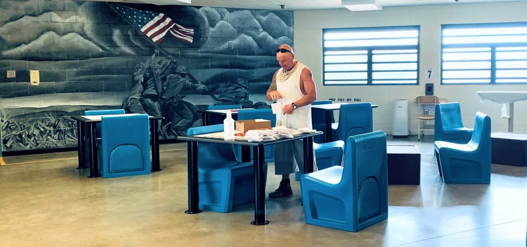 A man in a white tank top organizes items atop a table with blue chairs