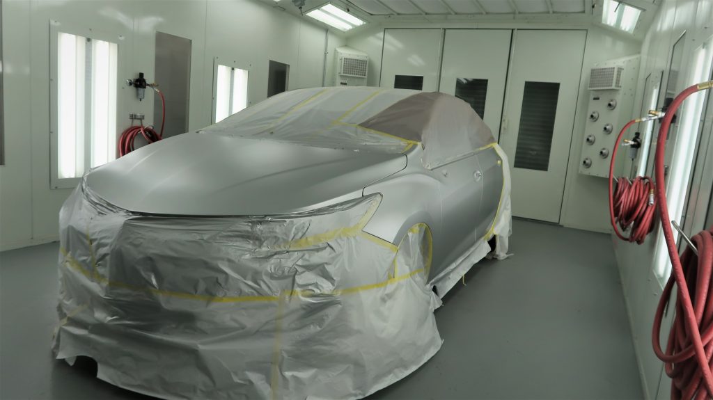 A silver car in a paint shop. The car is partially covered in painter's tape and protective material.