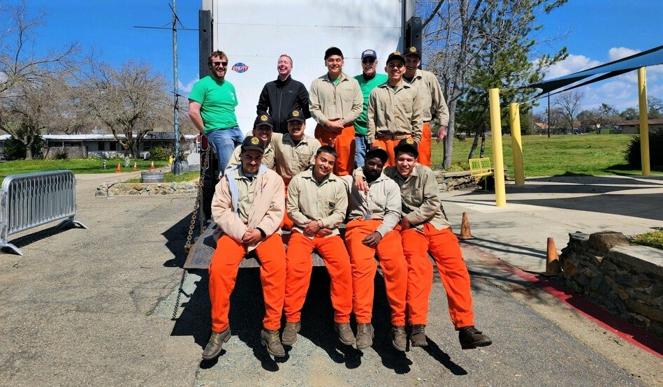 A group of young men in orange pants and tan shirts sit on the loading bed of a large truck. They are joined by two people in green shirts and one in a black coat, who is laughing.