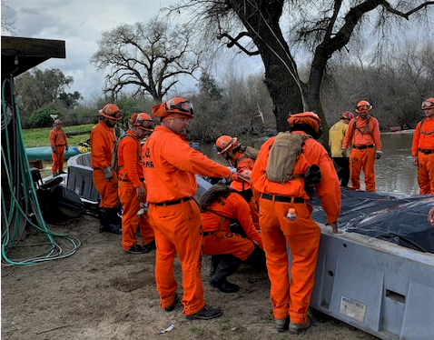 A large group of people in orange jumpsuits work on a retaining wall. A body of water is in the background.
