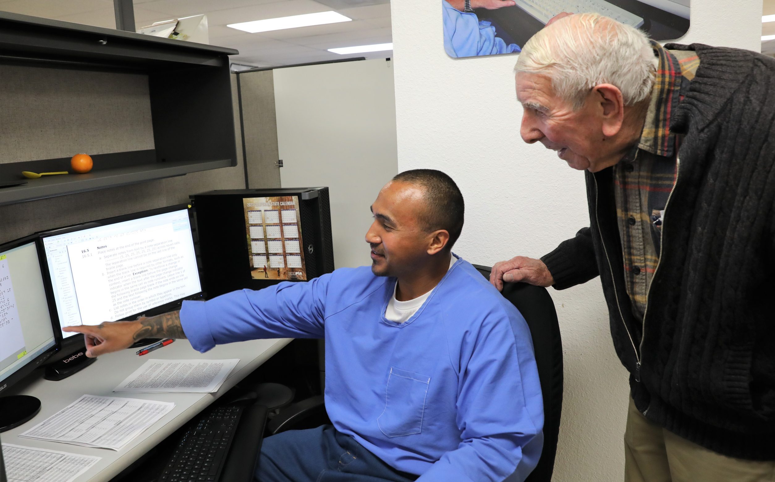 Elderly man looks over shoulder of CALPIA braille program participant at Folsom State Prison.