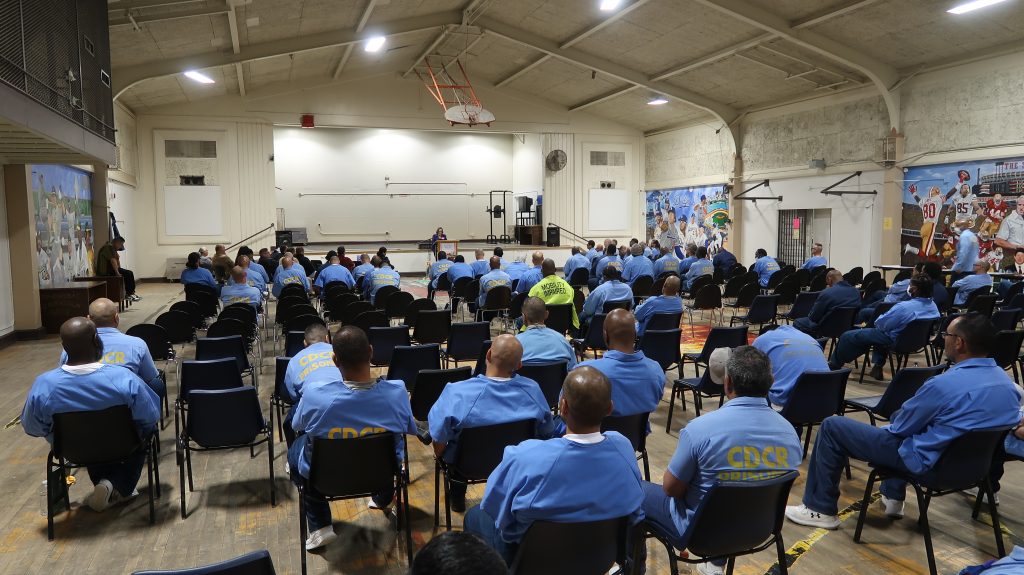 Incarcerated men sit in a gymnasium listening to a guest speaker.