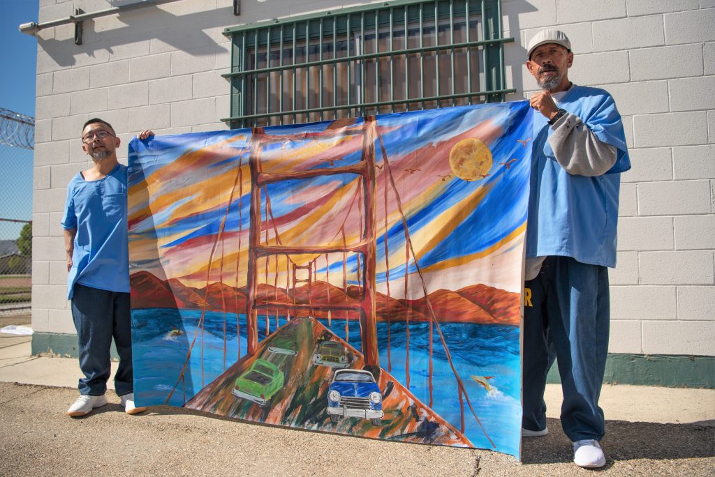 Two incarcerated artists hold a painting of the Golden Gate Bridge in San Francisco.