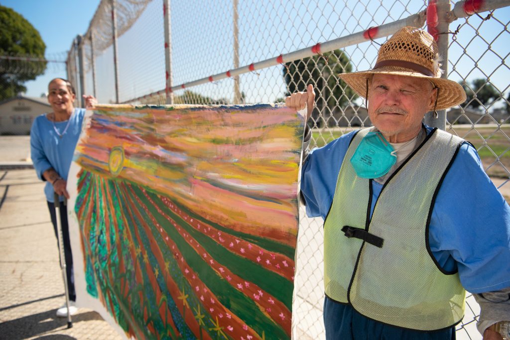 Two incarcerated artists hold a painting of a field of crops.