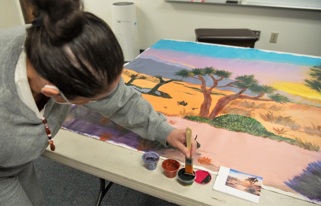Incarcerated female artist works on a landscape painting of Joshua trees and desert.