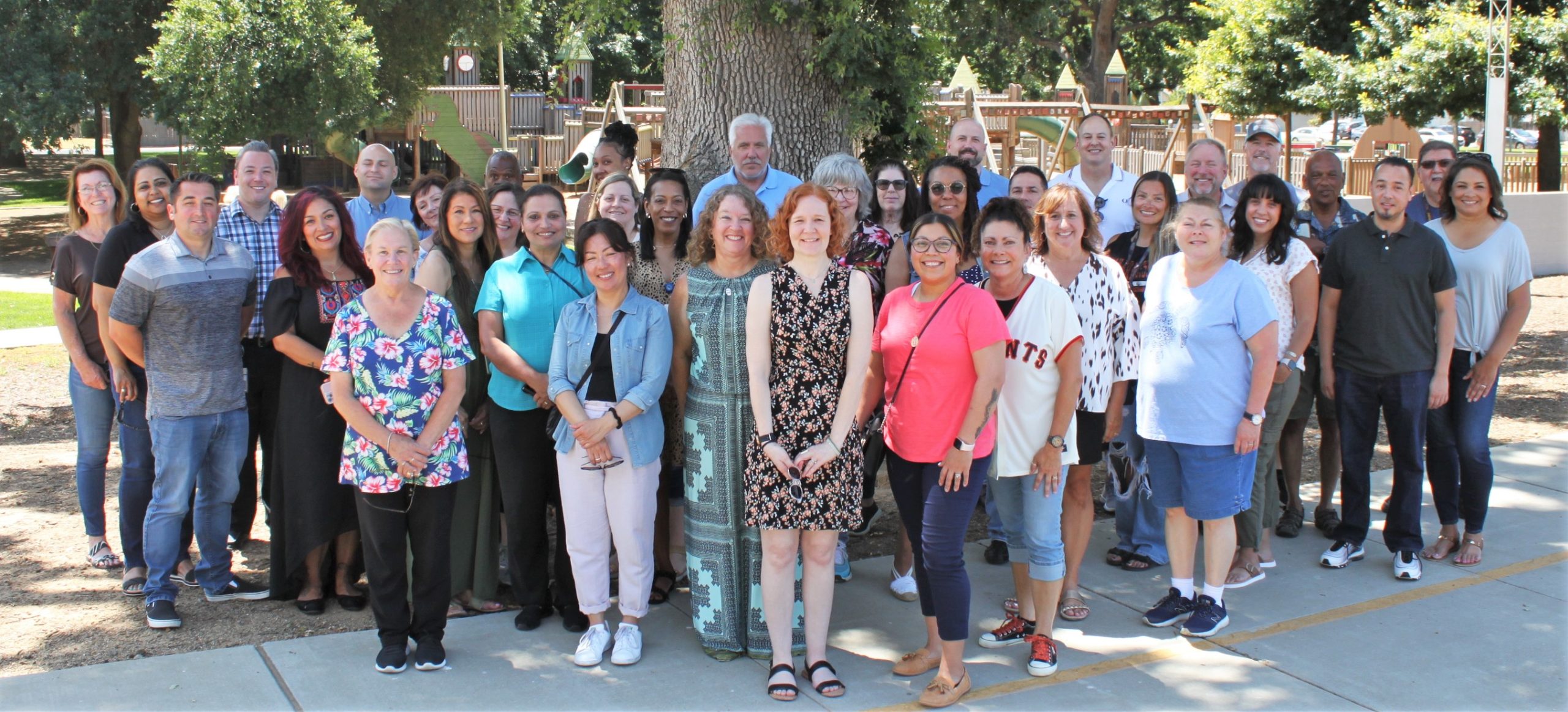 Current and former staff stand under a tree to pose for a group photo.