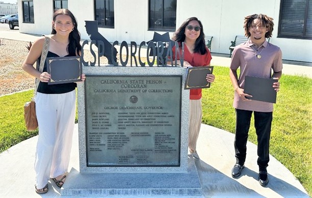 Three students hold certificates while standing beside the CSP-Corcoran plaque.