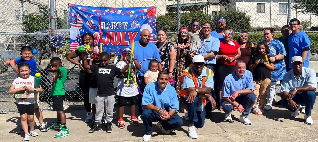 Group photo of incarcerated people with their visiting families taken in front of a fourth of july banner.