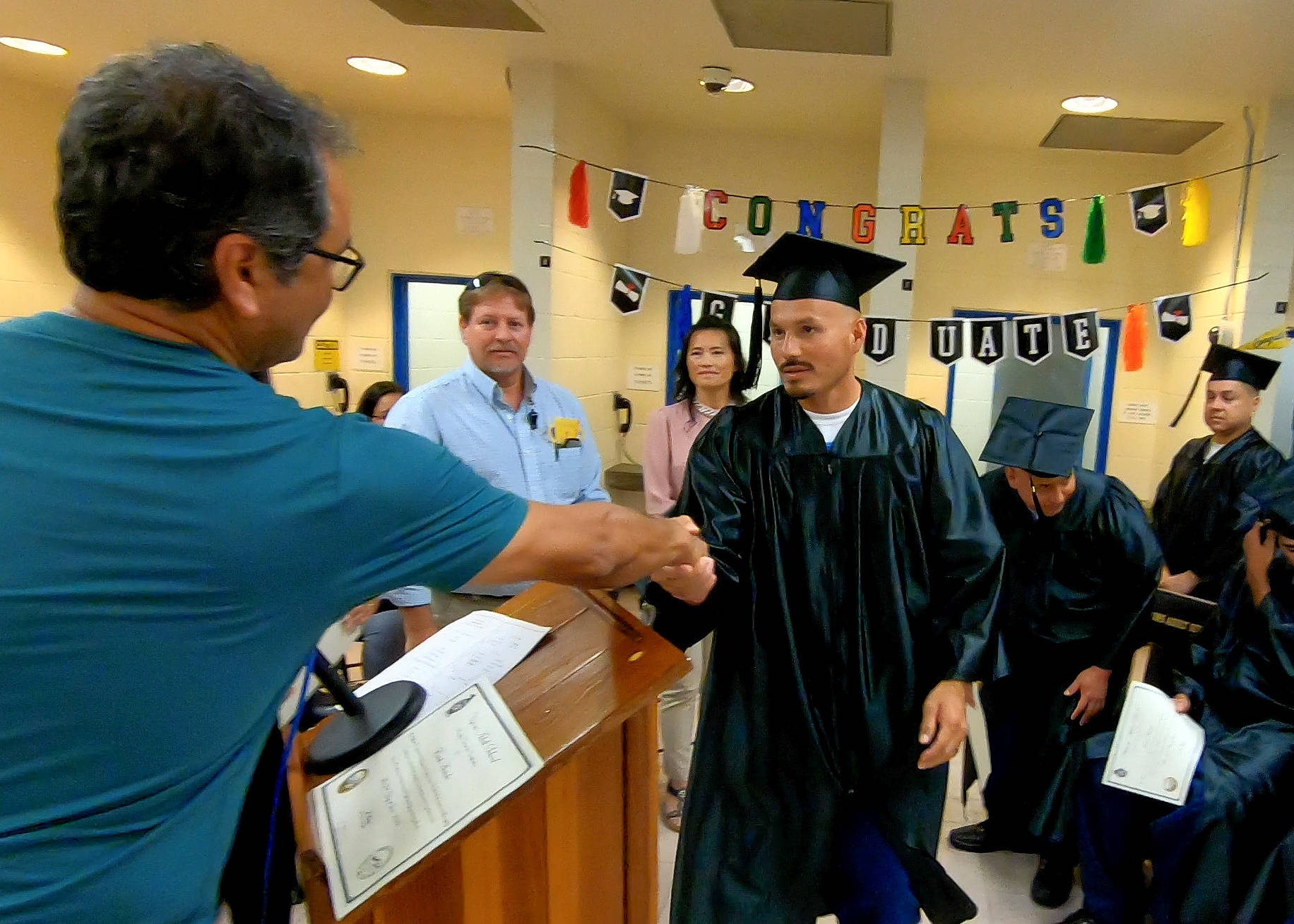 Graduations at COR for students earning college degrees and high school equivalency diplomas. Here a man shakes the hand of an incarcerated person wearing a cap and gown.