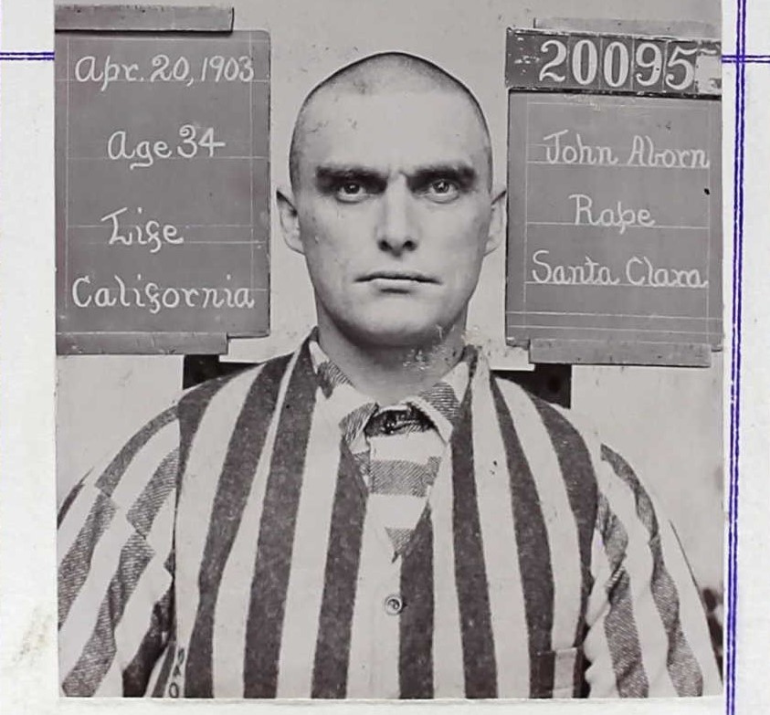 Mugshot of John Aborn wearing prison stripes in 1903. The information placards he's sitting between show the following information: Age 34, 20095, Rape, Santa Clara, Life sentence, California native.