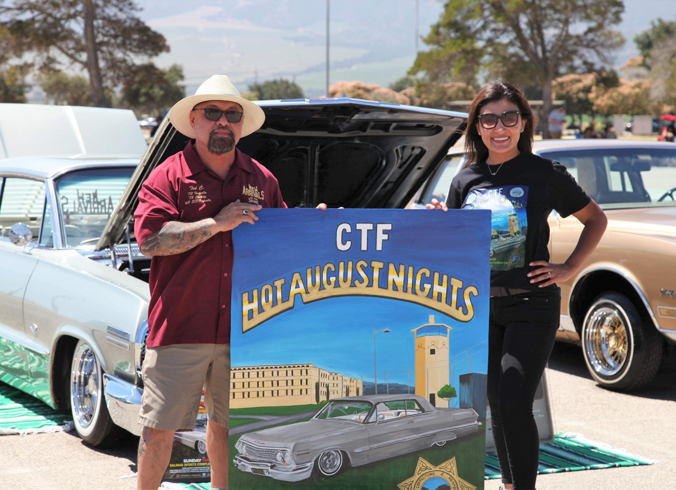 Two people at CTF hold a Hot August Nights car show sign while standing in front of vehicles.