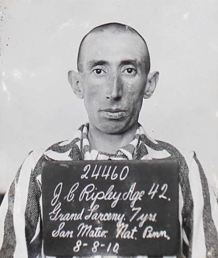 Man wearing prison stripes in 1910 at San Quentin. Prison intake board reads 24460, JC Ripley, 42, Grand Larceny, 7 years, San Mateo, Native of Pennsylvania.