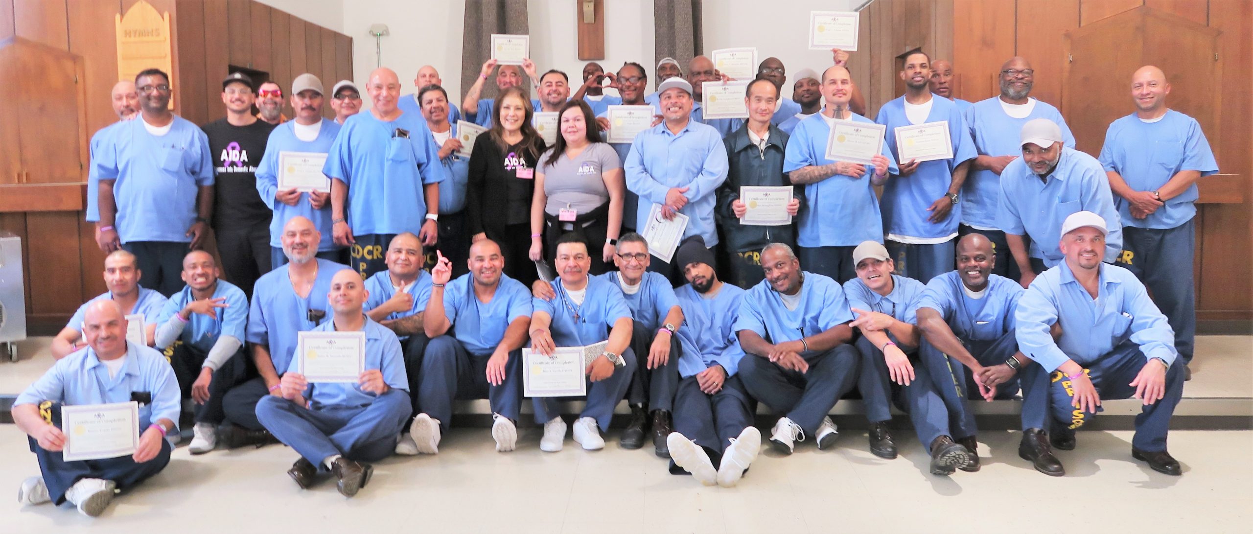 Incarcerated graduates of a domestic abuse program at Correctional Training Facility hold certificates, joined by program facilitators and volunteers.